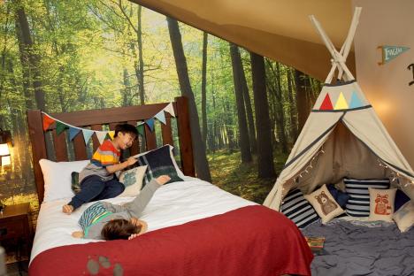 Two boys playing on a bed in a room decorated with a forest mural and a teepee.