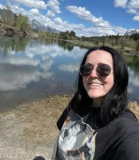 Woman wearing sunglasses smiles by a lake with mountains in the background.