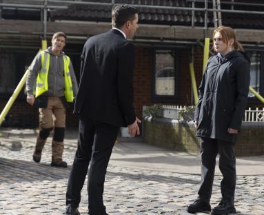 A man in a suit confronts a young woman, while a man in a high-visibility vest looks on.