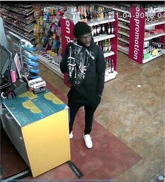A man in a black hat, black hoodie, and black pants stands in a store holding a black plastic bag.