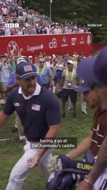 A male golfer in a US team shirt and hat walking past the camera while spectators cheer in the background.
