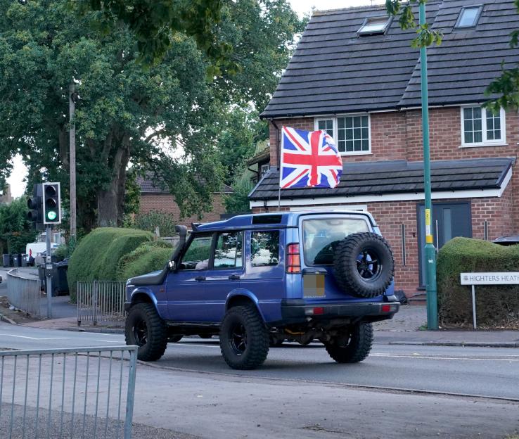 Blue Land Rover Discovery with a UK flag on its roof driving down a residential street.