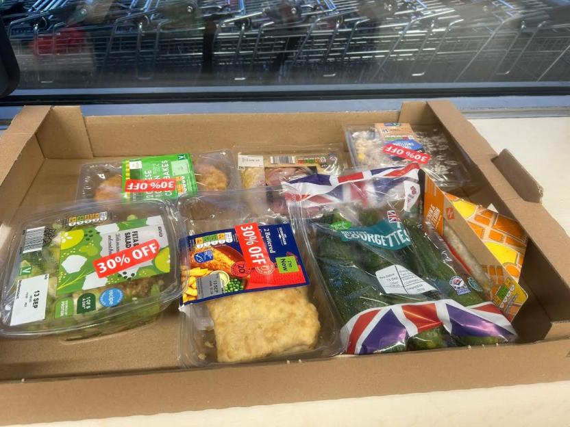Cardboard box filled with discounted food items, including salads, baked goods, and vegetables with "30% OFF" stickers.