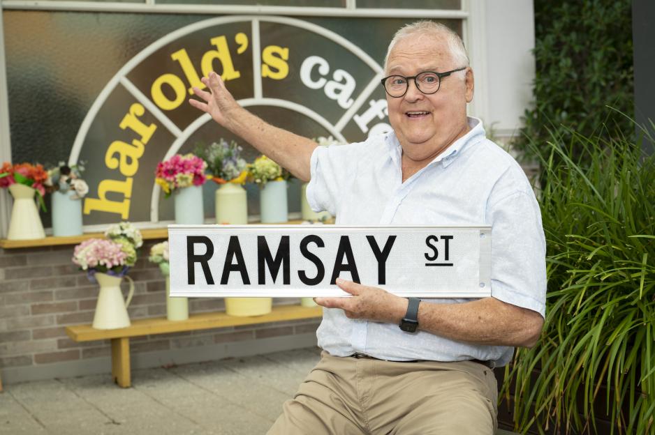 Ian Smith, holding a Ramsay Street sign, in front of Harold's Cafe.