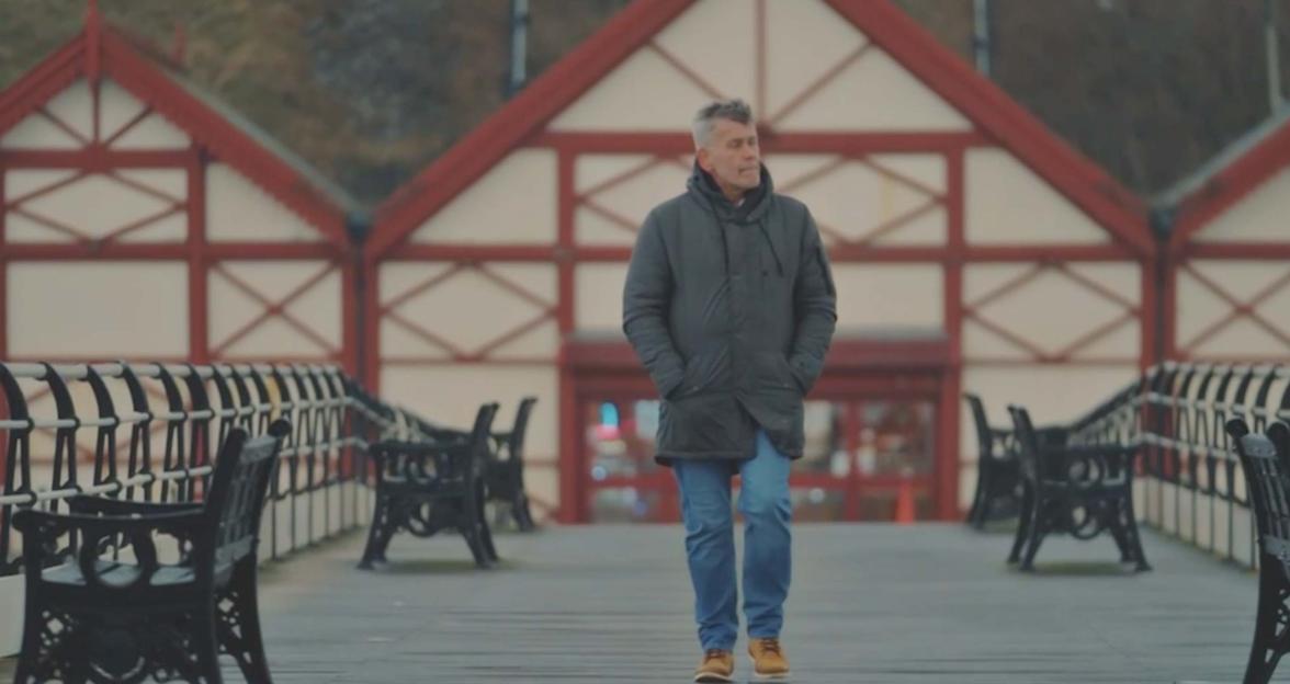 Paul, a groom from MAFS UK, walking along a pier with red and white buildings in the background.