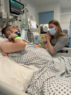 A woman in a face mask holds the hand of a man lying in a hospital bed.