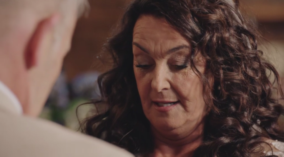 A middle-aged woman with dark, curly hair looks down, speaking softly to someone out of frame.