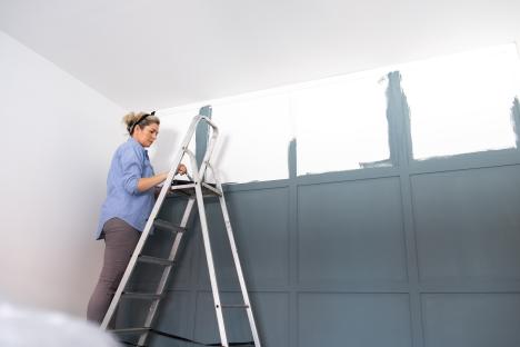 A woman on a ladder painting a paneled wall blue.