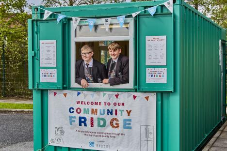 Two students stand in the window of a teal community fridge.