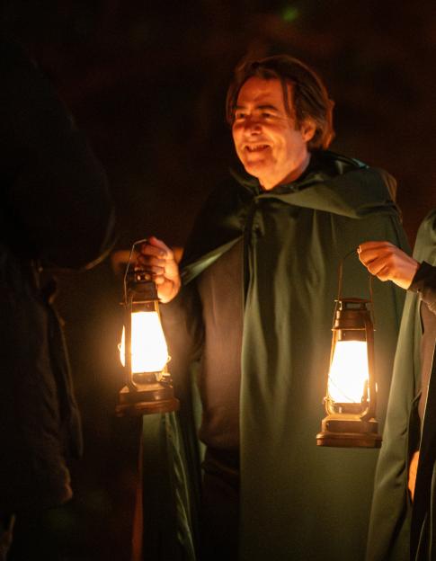 Three contestants from The Celebrity Traitors wearing dark green cloaks and holding lanterns.