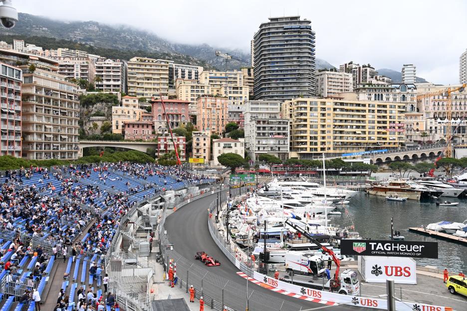 A red F1 car on the Circuit de Monaco race track, with spectators in bleachers and a harbor with yachts in the background, all set against a backdrop of mountainous city buildings.