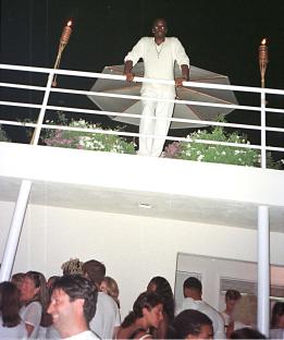 A man in white clothing stands on a balcony overlooking a party.