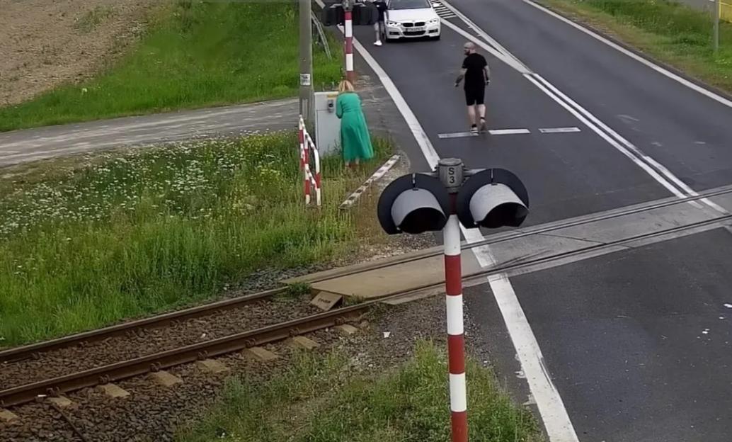 Security camera footage of a car crashing through a railway barrier and stopping on the tracks before being hit by a train.