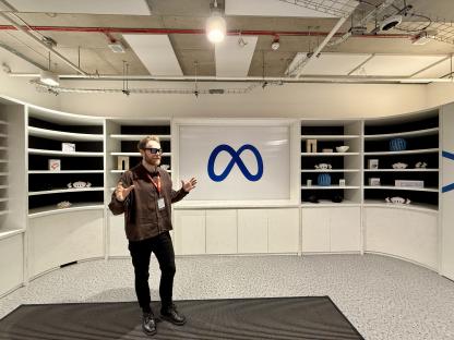 Man in glasses gesturing in front of a Meta logo displayed on a wall with shelves.