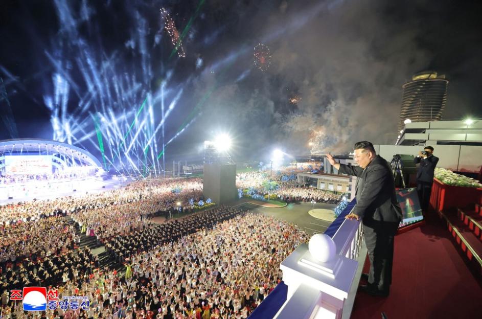 Kim Jong-un waving to a large crowd at a nighttime event with fireworks.