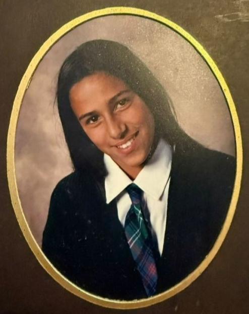 Photo of Correne Dadd from a yearbook, showing her with long dark hair, a white shirt, a plaid tie, and a dark jacket, smiling at the camera.
