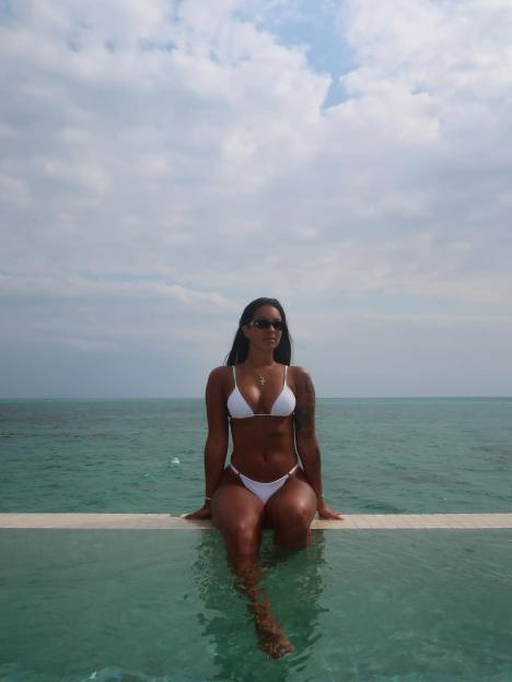 A woman in a white bikini and sunglasses sits on the edge of an infinity pool with the ocean in the background.
