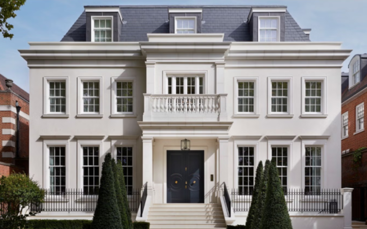 Exterior view of a large white stucco house with a dark gray slate roof and a black double door, flanked by evergreen trees and a black metal fence.