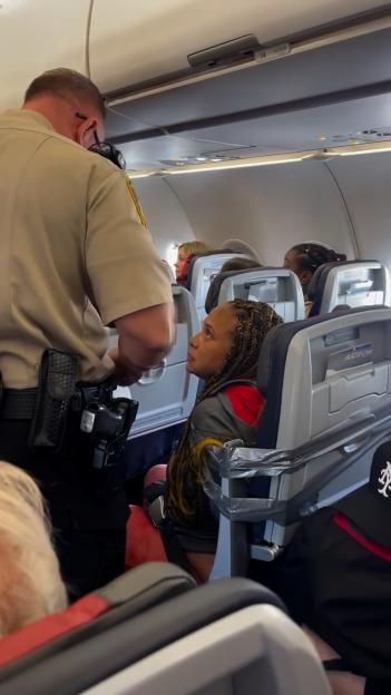 A passenger restrained with duct tape in an airplane seat, looking up at an officer.