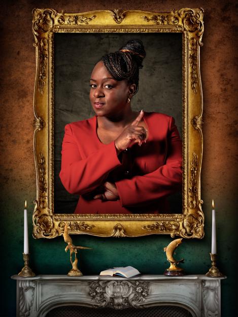 Tameka Empson in a red blazer, framed within a gilded mirror above a fireplace with lit candles, two golden figurines, and an open book.