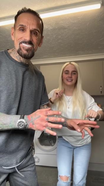 A man with a beard and tattoos on his arms and neck, wearing a gray shirt, smiles at the camera while dancing next to a woman with long blonde hair, wearing a white shirt and light-blue ripped jeans, who is also dancing.