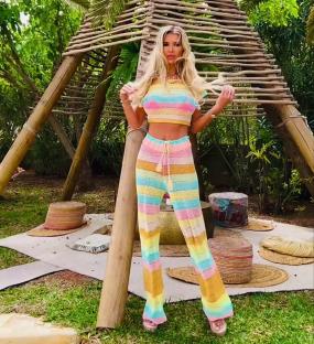 Woman in rainbow striped knit pants and top standing near a wooden structure.