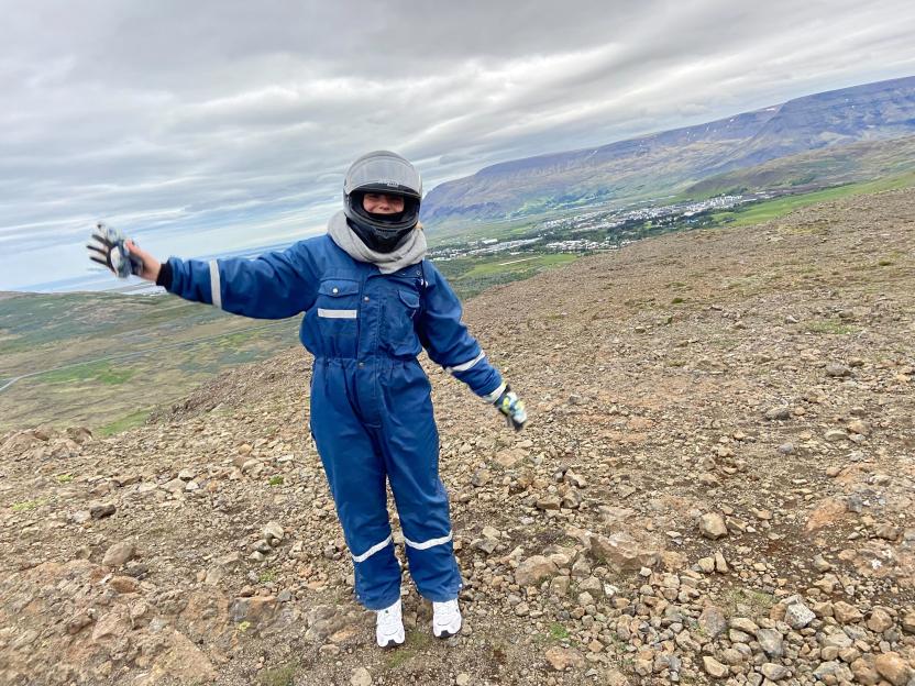 A person in a blue jumpsuit, helmet, and gloves stands on a rocky hill overlooking a town and mountains.
