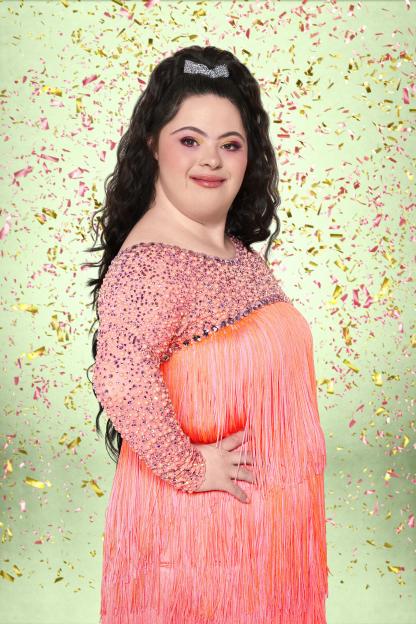 Ellie Goldstein with long dark hair, wearing a sequined pink and orange fringed dress, standing against a light green background with falling confetti.