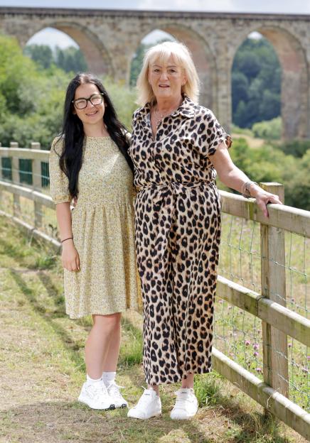 Two women stand together by a fence, a stone viaduct in the background.