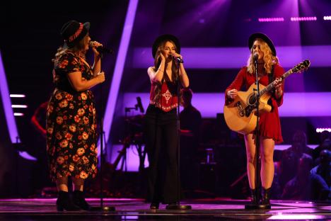 Three women singing on stage, one playing an acoustic guitar.