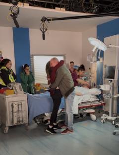Behind-the-scenes photo of a hospital scene; a man and woman embrace while others look on.