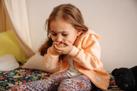 A young girl sitting on a bed, covering her mouth with her hands, appearing unwell.