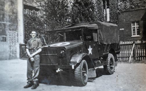 Black and white photo of a young man in uniform standing next to a military truck.