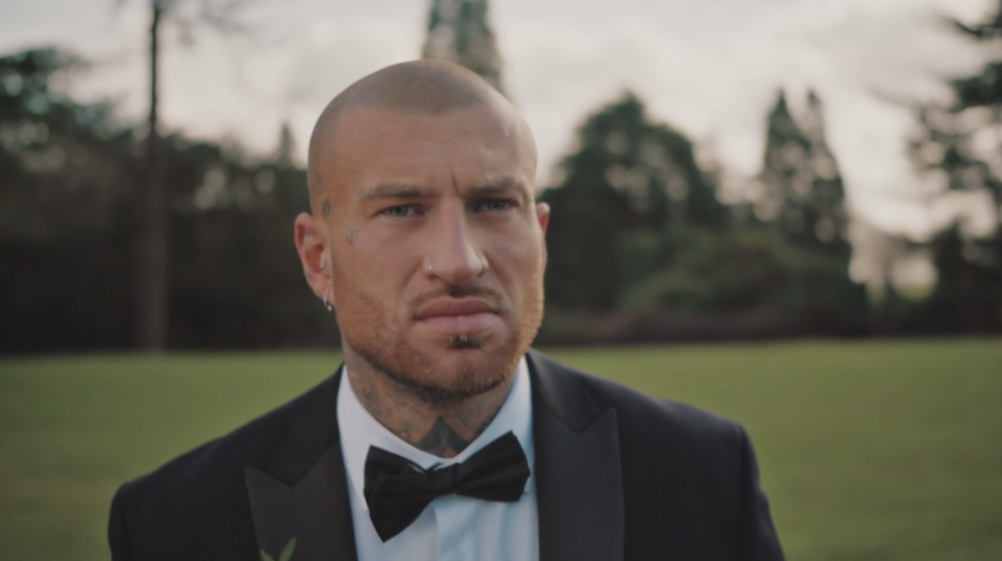 A man with a shaved head and tattoos on his face and neck wearing a tuxedo with a bow tie and a boutonnière.