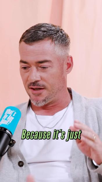 A man with short grey hair and a beard wearing a white t-shirt and grey cardigan speaks into a blue microphone.