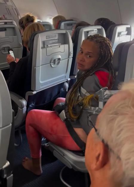 A woman taped to her airplane seat looks over her shoulder at the camera.