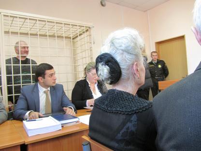 A courtroom scene showing a man in a cage, other individuals seated at a table, and a guard standing nearby.