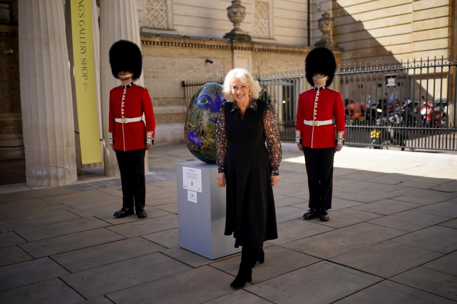 Queen Camilla standing by a large decorated egg sculpture outside Buckingham Palace, flanked by two guardsmen.