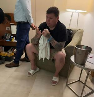 Man seated in a hotel room, holding a towel and looking down at his hands.