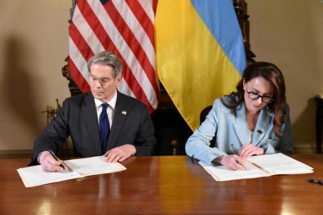Two people signing a document at a table with the American and Ukrainian flags in the background.