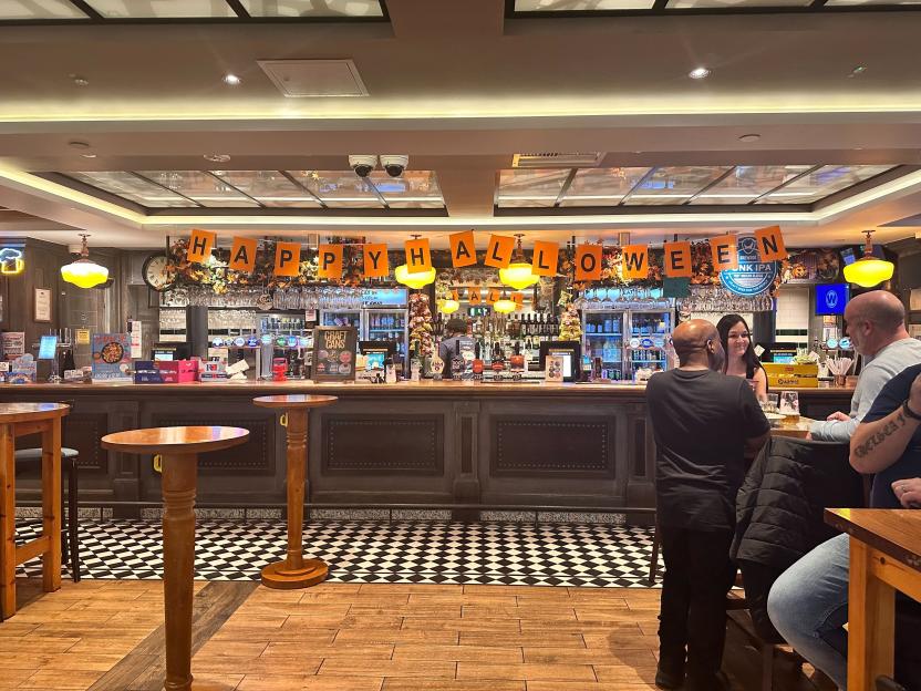 A pub interior decorated for Halloween, featuring a long bar with drinks and several patrons.
