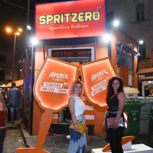 Two women stand in front of a Spritzero aperitivo italiano stand holding large Aperol glasses.