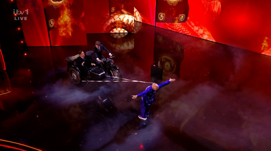 Screenshot of a man being pulled by a motorcycle sidecar on a stage.