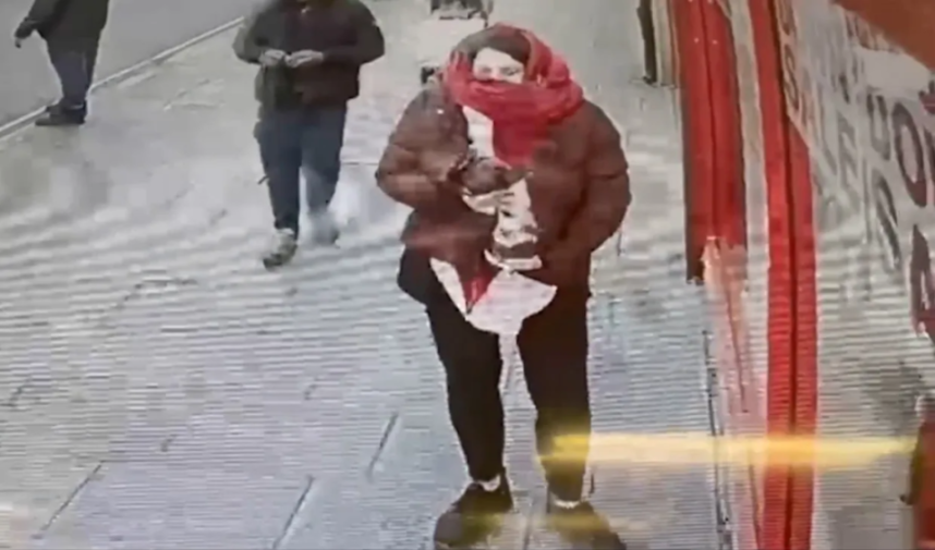 A person in a red scarf and brown jacket walking on a sidewalk.