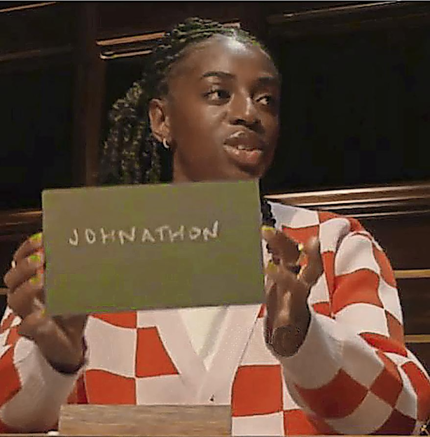 A woman in a red and white checkered sweater holds a green sign that reads "JOHNATHON" in white letters.