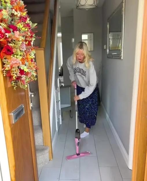 A woman in a long skirt and sweatshirt mops a light-colored tile floor in a hallway.