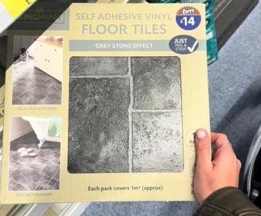 Person holding a box of grey stone effect self-adhesive vinyl floor tiles.