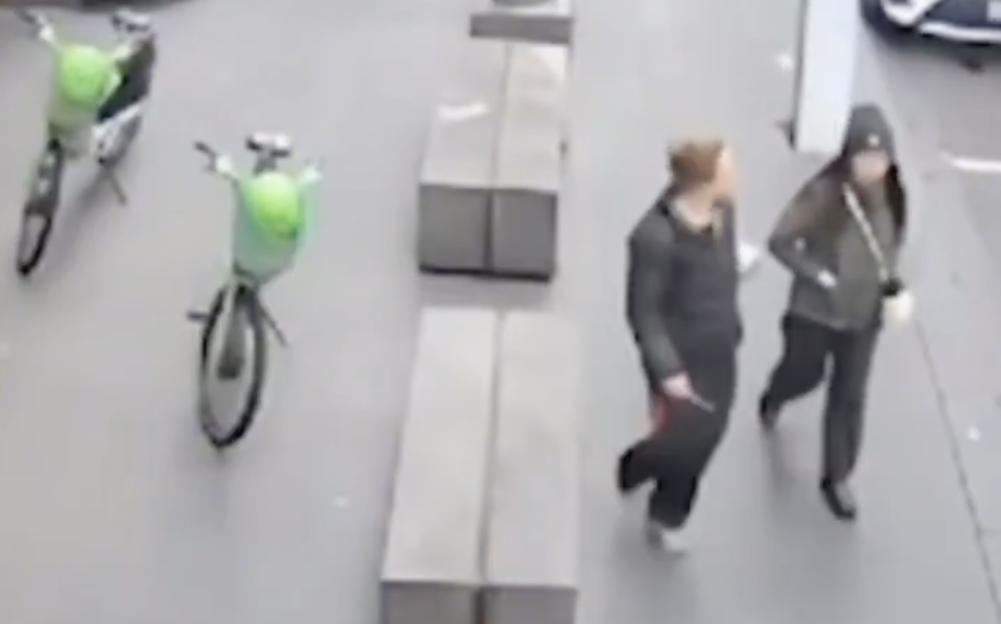Two people walking on a sidewalk with two bicycles and several concrete benches.