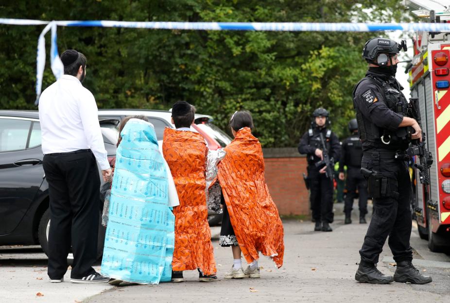Witnesses and victims, some wrapped in emergency blankets, at a crime scene near a synagogue where police are present.