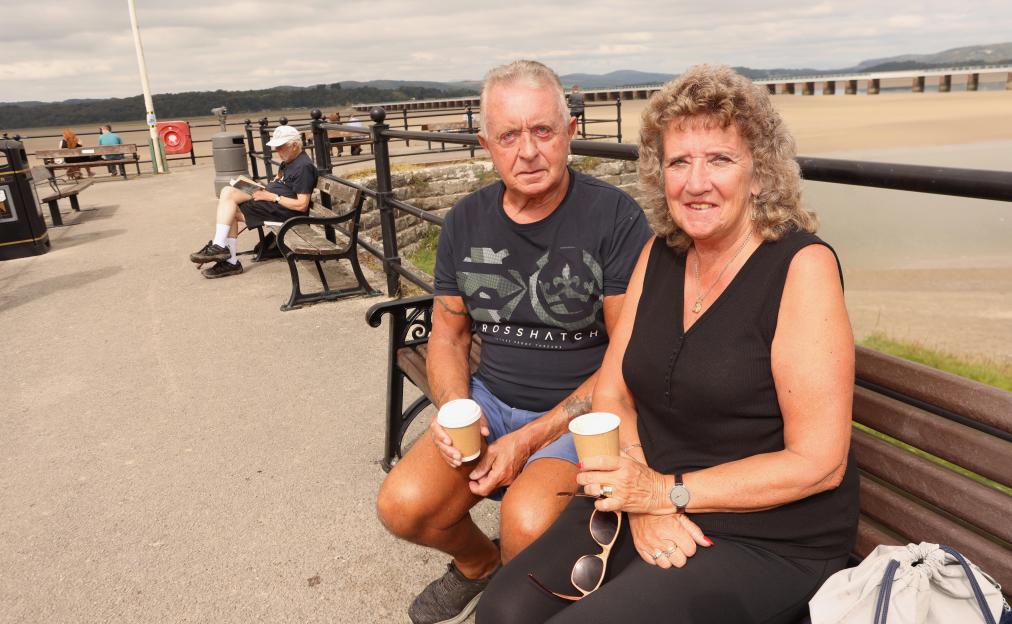 A couple sits on a park bench by the water, holding takeaway coffee cups.
